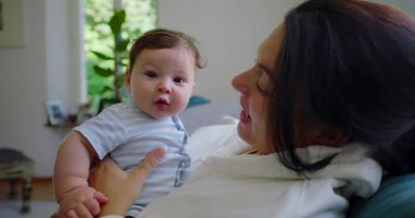 Bebek doğrudan kameraya bakarken anne kucağında yumuşak doğal ışık ve huzurlu bir ev atmosferiyle aydınlık bir oturma odasında neşeli ve hassas bir an yaratıyor.