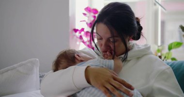 Anne, bebeğini nazikçe kucağına alıp bir kanepede kucaklar ve sıcak bir yuva ortamında sevgi ve özenle dolu huzurlu ve şefkatli bir an yaratır.