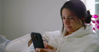 Anne kanepeye oturuyor, bebeğini kucağında tutuyor ve ev ortamında odaklanmış bir ifadeyle telefonuna bakıyor.