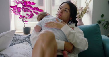 Anne, bebeğini nazikçe kucağına alıp bir kanepede kucaklar ve sıcak bir yuva ortamında sevgi ve özenle dolu huzurlu ve şefkatli bir an yaratır.