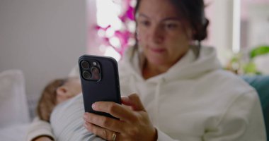 Yavrusu huzurlu bir oturma odasında, parlak doğal ışıkla kucağında dinlenirken bir annenin telefonunu kullanışının yakın çekimi.