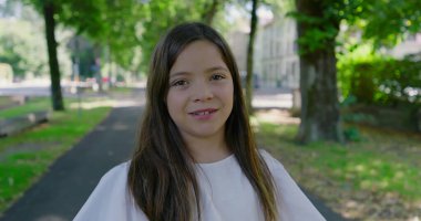 Parkta ağaçlarla kaplı bir yolda duran genç kız, kameraya tarafsız bir ifadeyle bakıyor, yeşil ve güneş ışığıyla çevrili, sakin bir açık hava anını yakalıyor.