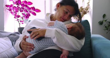 Anne bebeğini kollarında tutarken sıcak bir şekilde gülümsüyor. Yumuşak ışıkla sıcak bir ev ortamında şefkatli ve şefkatli bir an yaratıyor.