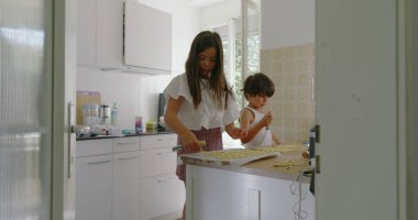 Kardeşler mutfakta birlikte yemek hazırlıyorlar, kız hamura odaklanıyor, oğlan asistanlığı, aile yemekleri, parlak mutfak, takım çalışması, anne arka planda, çekici bir an.