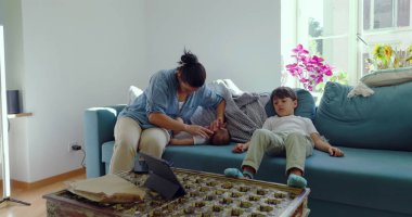 Annesi bebeğiyle ilgilenirken, küçük oğlu da penceresinden içeri ışık saçan parlak bir oturma odasındaki kanepede oturuyor.