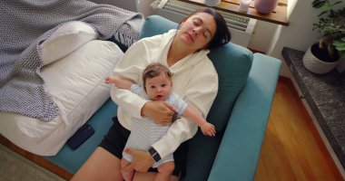 Anne, bebek göğsünde tutarken gözleri kapalı dinleniyor. Aydınlık ve rahat bir ev ortamında kanepede sakin ve huzurlu bir anın tadını çıkarıyor..