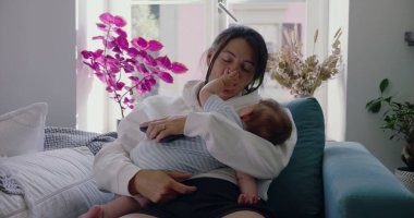 Anne bebeğini kucağında, bebek yüzüne dokunurken yavaşça kanepede dinleniyor. Huzurlu bir ev ortamında samimi bir yakınlık ve bağlantı anı. SD 480p