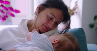 Anne bebeğini kucaklarken onlara sevgiyle gülümsüyor, güneşli bir oturma odasında huzurlu ve besleyici bir an, anne sıcaklığı ve şefkat gösteriyor.