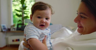Bebeğin annesinin kollarındayken kameraya baktığı yakın plan, bebeğin parlak, ev ortamındaki meraklı ve nişanlı ifadesi..