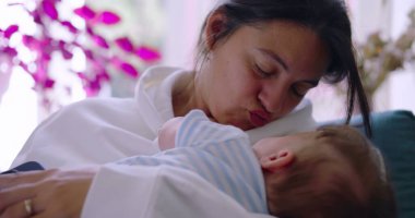 Anne bebeğini kucaklarken onlara sevgiyle gülümsüyor, güneşli bir oturma odasında huzurlu ve besleyici bir an, anne sıcaklığı ve şefkat gösteriyor.