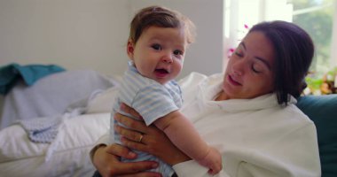 Bebek doğrudan kameraya bakarken anne kucağında yumuşak doğal ışık ve huzurlu bir ev atmosferiyle aydınlık bir oturma odasında neşeli ve hassas bir an yaratıyor.