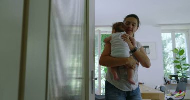 Anne bebeğini kucağında bir ev koridorunda yürürken gerçek anne ve aile hayatının özgün bir anını tasvir ediyor.