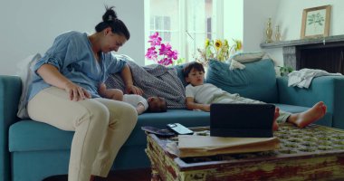 Annesi bebeğiyle ilgilenirken, küçük oğlu da penceresinden içeri ışık saçan parlak bir oturma odasındaki kanepede oturuyor.