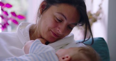 Anne bebeğini kucaklarken onlara sevgiyle gülümsüyor, güneşli bir oturma odasında huzurlu ve besleyici bir an, anne sıcaklığı ve şefkat gösteriyor.
