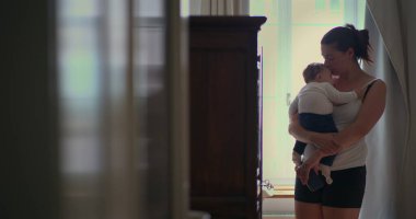 Anne bebeğini sevgi dolu bir şekilde kucağında tutuyor. Yumuşak ışıklı bir odada yürürken huzurlu bir bağ anı yaratıyor ve sıcak bir yuva kuruyor.