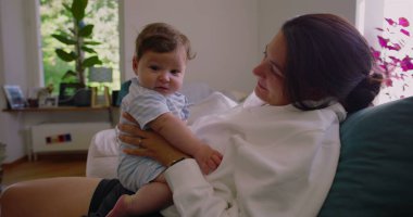 Anne, güneşli bir oturma odasında bebeğine gülümserken anne ve çocuk arasında sıcak ve sevgi dolu bir bağ kuruyor.
