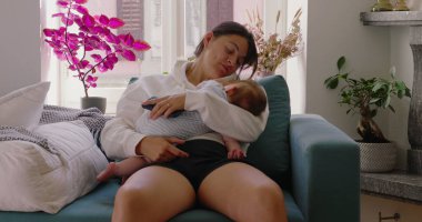 Anne bebeğini rahat bir pozisyonda, rahat bir oturma odasında koltukta otururken tutuyor, anne ve çocuk arasında sakin ve şefkatli bir an.