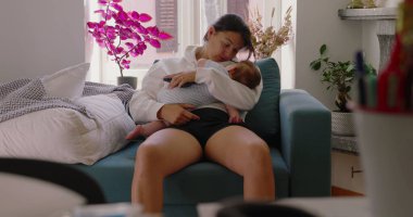 Anne bebeğini rahat bir pozisyonda, rahat bir oturma odasında koltukta otururken tutuyor, anne ve çocuk arasında sakin ve şefkatli bir an.