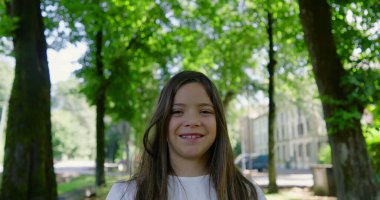 Güneşli bir parkta ağaçlarla kaplı bir patikada duran uzun saçlı, gülümseyen kız beyaz bir bluz giyiyor, yeşilliğin ve doğanın tadını çıkarıyor, arka planda binalar var.