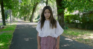 Düşünceli bir kız, sakin, ağaçlarla kaplı bir parkta tek başına dikiliyor, kameraya bakıyor, günlük beyaz bir bluz ve desenli bir etek giyiyor.