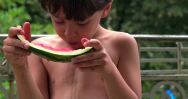Genç çocuk dikkatlice bir dilim karpuz yiyor. Güneşli bir günde taze ve tatlı meyvenin tadını çıkarırken meyve suyu damlatıyor. Anın tadını çıkarıyor.