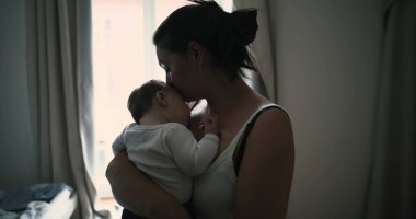 Anne pencerenin kenarında duruyor, bebeği kucaklıyor ve sevgi ve sevgiyle aşağı bakıyor, evde huzurlu ve samimi bir aile anı yakalıyor.