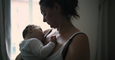 Anne pencerenin kenarında duruyor, bebeği kucaklıyor ve sevgi ve sevgiyle aşağı bakıyor, evde huzurlu ve samimi bir aile anı yakalıyor.