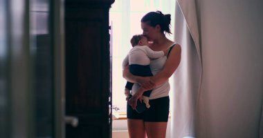 Anne bebeğini hafifçe aydınlatılmış bir odada kucağında tutuyor, şefkatli bir ilgi ve bağlantı anı sergiliyor, sakin bir ortamda sevgiyi ve bakımı ön plana çıkarıyor.