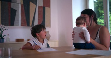 Masada evde eğitim görürken gözünü ovuşturan yorgun çocuk, yanında bebeğini kucağında tutan anne, aile dinamiklerini ve eğitim odaklarını ön plana çıkarıyor.