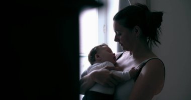 Anne, bebeğini nazikçe bir pencerenin yanında tutarak evdeki yumuşak doğal ışıkta sessiz bir bağ anını paylaşıyor.