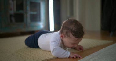 Bebek karnının üstünde, yerden yukarı bakıyor, merakını ve hayal kırıklığını ifade ediyor, rahat bir ev ortamında emekleme ve keşiflerin ilk aşamalarında yer alıyor.