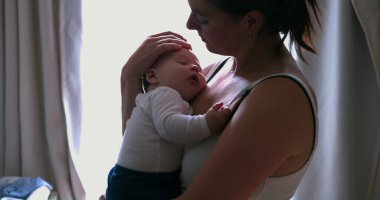 Bir annenin, uyuyan bebeğini kucakladığı, sessiz bir evde sevgi ve güvenlik sağladığı, pencerenin ışıklarıyla hafifçe aydınlattığı o hassas an.