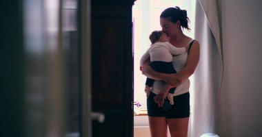 Anne bebeğini hafifçe aydınlatılmış bir odada kucağında tutuyor, şefkatli bir ilgi ve bağlantı anı sergiliyor, sakin bir ortamda sevgiyi ve bakımı ön plana çıkarıyor.