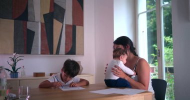 Masada evde eğitim görürken gözünü ovuşturan yorgun çocuk, yanında bebeğini kucağında tutan anne, aile dinamiklerini ve eğitim odaklarını ön plana çıkarıyor.