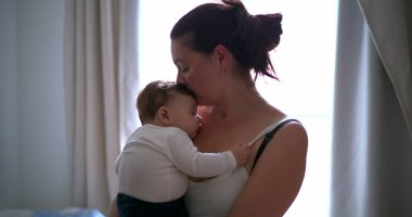 Anne bebeğini kucağında tutuyor, bebeği huzurlu bir ortamda nazikçe kucaklıyor ve rahatlatıyor, sıcaklığı ve bağlantıyı vurguluyor.