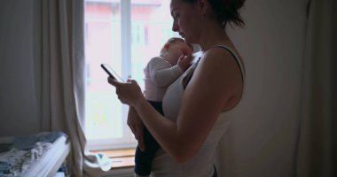 Anne bebeğini akıllı telefon kullanırken kucağında tutuyor, penceresinden gelen yumuşak doğal ışıkla ev ortamında birden fazla iş yapıyor.