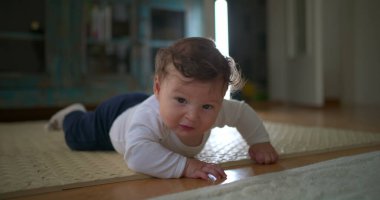 Bebek karnının üstünde, yerden yukarı bakıyor, merakını ve hayal kırıklığını ifade ediyor, rahat bir ev ortamında emekleme ve keşiflerin ilk aşamalarında yer alıyor.