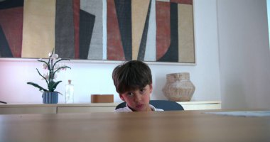 Genç Çocuk üzgün ve kızgın bir ifadeyle masada oturuyor, sakin bir ev ortamında hayal kırıklığı yaşıyor, aile ortamında duygular ve gerilim hissediyor.