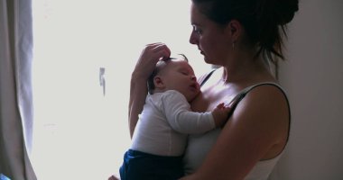 Bir annenin, uyuyan bebeğini kucakladığı, sessiz bir evde sevgi ve güvenlik sağladığı, pencerenin ışıklarıyla hafifçe aydınlattığı o hassas an.