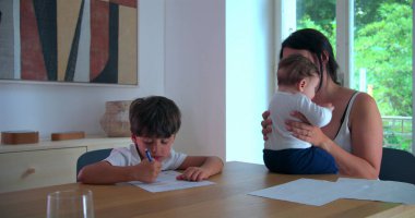 Çocuk, anne yanında bebeği tutarken evde eğitim işine odaklanmış, aile desteğini göstermiş ve sakin bir evde eğitim ve büyüme ortamı sağlamıştır.