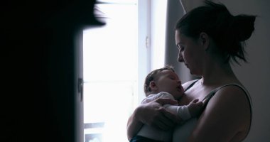 Anne, bebeğini nazikçe bir pencerenin yanında tutarak evdeki yumuşak doğal ışıkta sessiz bir bağ anını paylaşıyor.