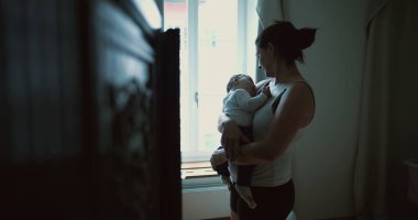 Anne pencerenin kenarında duruyor, bebeği kucaklıyor ve sevgi ve sevgiyle aşağı bakıyor, evde huzurlu ve samimi bir aile anı yakalıyor.