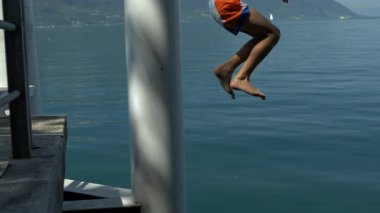 Çocuk rıhtımdan yavaş çekime atlıyor, bacakları açık mavi suyun üzerine doğru uzanıyor, neşeli yaz macerası ve kaygısız çocukluk enerjisi.