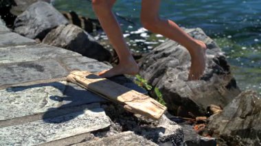 Çıplak ayaklı çocuk göl kıyısında kayalık yolda yürüyor, yavaş çekim videosu doğal hareketleri, açık hava enerjisini ve su kenarında kaygısız yaz keşiflerini gösteriyor.