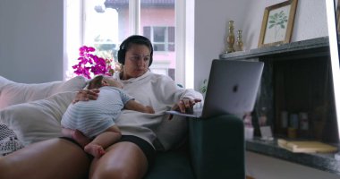 Anne dizüstü bilgisayarda çalışırken bebek göğsünde uyuyor kulaklık takıyor, rahat oturma odası, iş ve ebeveynlik dengesi, yumuşak doğal ışık.