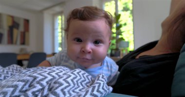 Bebek battaniyeye uzanmış merakla bakarken anne arka planda doğal ışık ve sıcak aile atmosferiyle dolu rahat bir oturma odasında gülümsüyor.