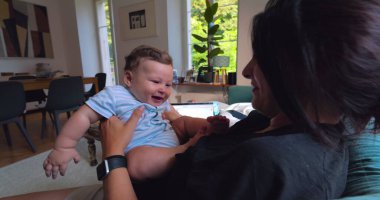 Anne, anne ve çocuk arasındaki bağ ve sevgi dolu doğal ışıkla dolu parlak bir oturma odasında oynarken, gülümserken ve gülerken bebeği havaya kaldırıyor.