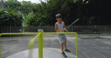 Genç bir çocuk oyun parkında enerjik bir şekilde koşuyor. Atlıkarınca, sıcak bir günde dışarıda oyun oynuyor. Etrafı yemyeşil ve park ekipmanlarıyla çevrili.