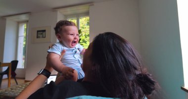 Anne, anne ve çocuk arasındaki bağ ve sevgi dolu doğal ışıkla dolu parlak bir oturma odasında oynarken, gülümserken ve gülerken bebeği havaya kaldırıyor.