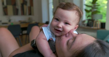 Anne, sevgi dolu ve sevgi dolu bir anda bebeği öpüyor ve kucaklıyor. Parlak bir oturma odasında oturuyor. Neşe ve yakınlık içinde kaynaşıyor.